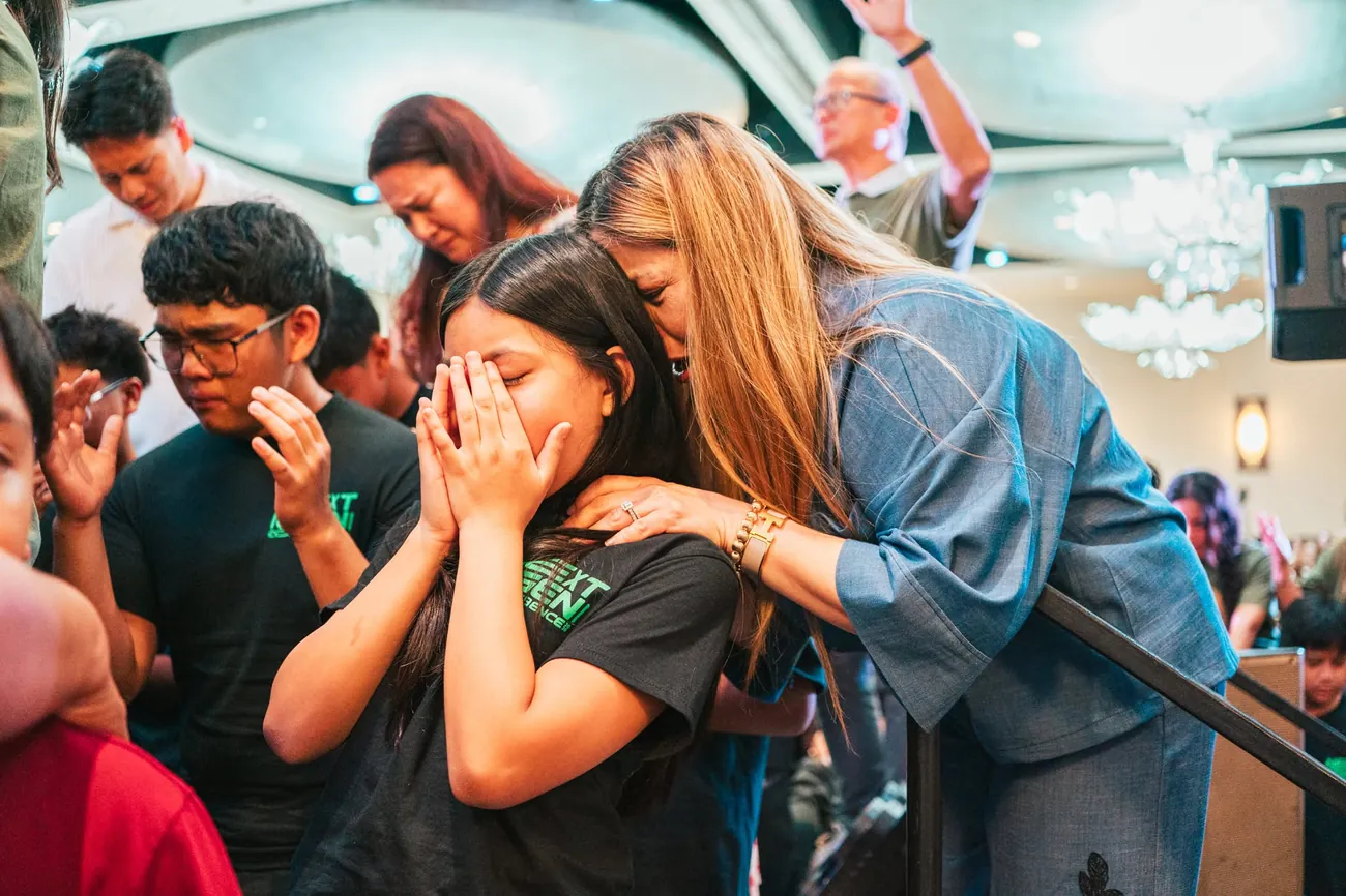 A woman comforting a young woman in prayer during worship, heads bowed together