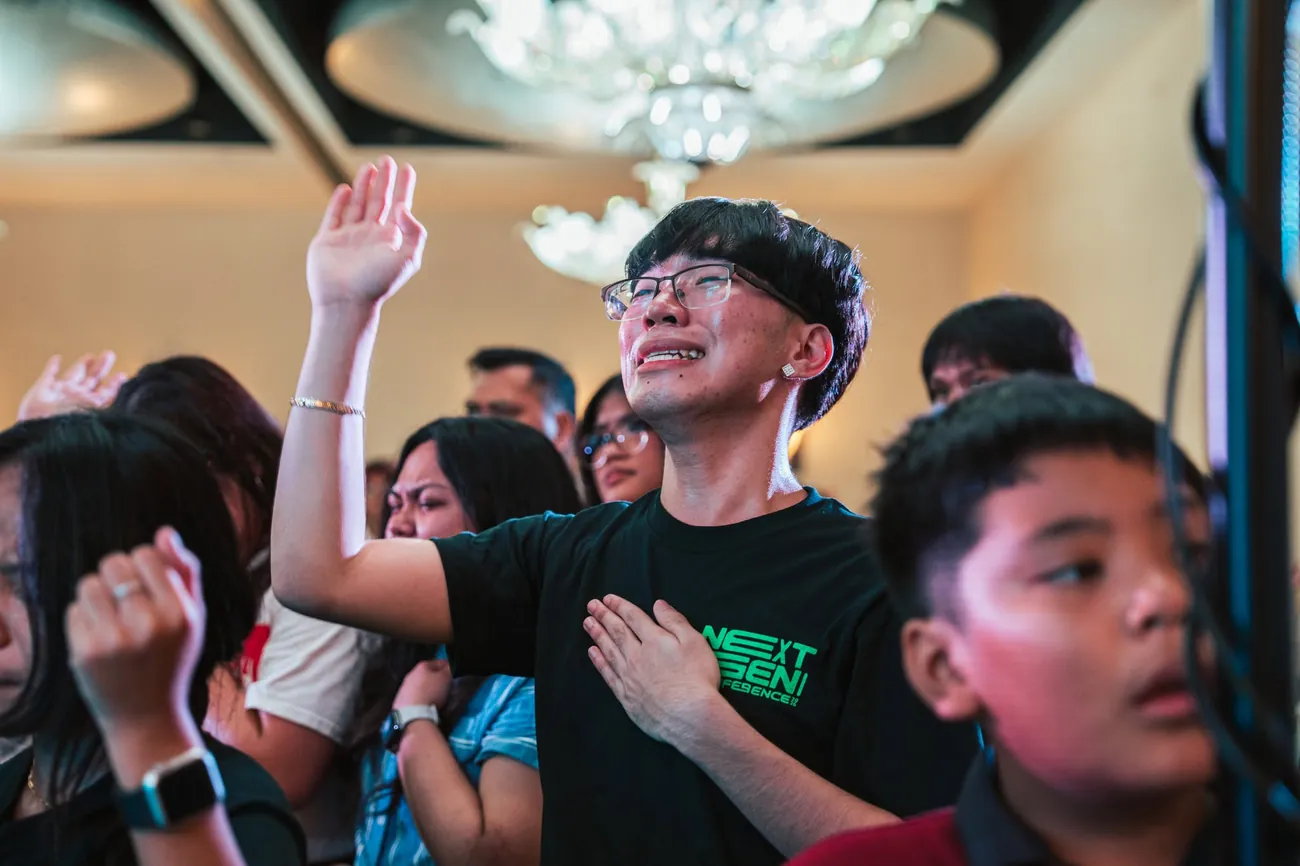 A young man praying with his hand raised, a chandelier glowing above