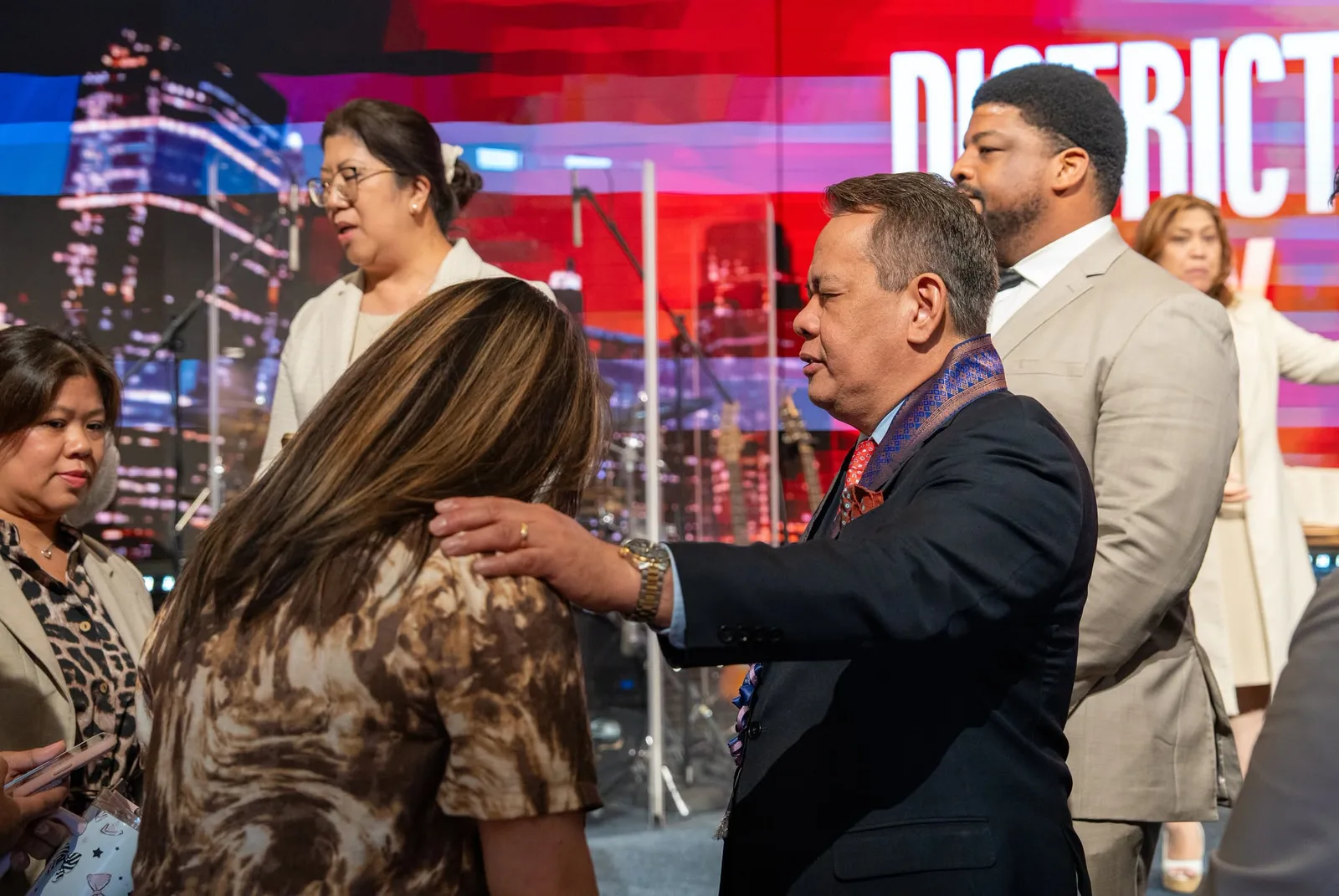 A minister welcoming a guest with a hand on her shoulder, the district LED wall glowing behind them
